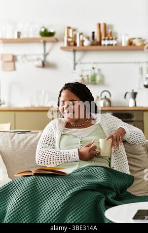 Una giovane donna gode di una mattinata tranquilla avvolta in una coperta mentre sorseggia tè e legge. Foto Stock