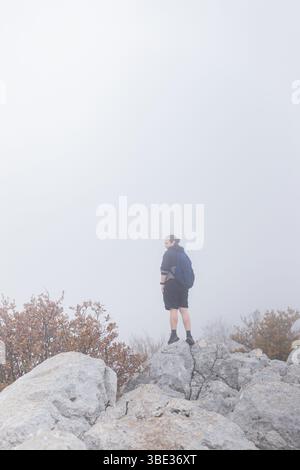 L'uomo si erge in cima alla cresta rocciosa delle montagne indossando un abbigliamento sportivo circondato da fitta nebbia e arbusti autunnali. Foto Stock