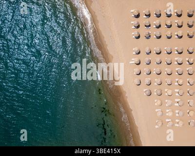 Vista aerea dall'alto verso il basso di una spiaggia sabbiosa con ombrelloni di paglia e lettini allineati in file, onde che lavano delicatamente a riva. Concetto di vacanza estiva Foto Stock