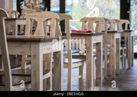 Uno spazio caffetteria o ristorante con diversi tavoli e sedie di legno chiaro disposti in file e una parete luminosa con finestre visibili sullo sfondo, creat Foto Stock