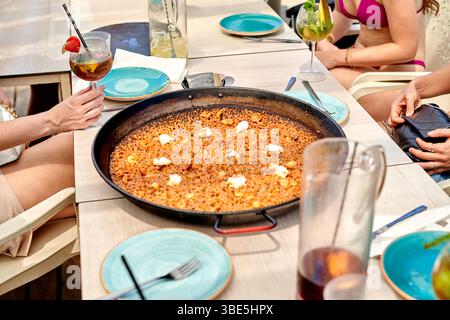 Gli amici si riuniscono intorno a un tavolo su una spiaggia, assaporando una grande paella di pesce accompagnata da bevande colorate. L'atmosfera e' vivace e vibrante con la Foto Stock