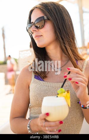 Una donna con gli occhiali da sole si gode un cocktail rinfrescante in una spiaggia soleggiata. Tiene una bevanda fruttata adornata con ananas e lime, vestita per il rela estivo Foto Stock