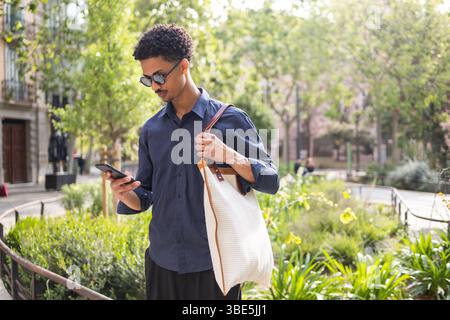 Un giovane in città, vestito con un abbigliamento casual, si è concentrato sul telefono mentre tiene in mano una borsa. La luce naturale illumina l'interno del punto vendita verde. Foto Stock