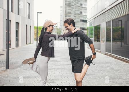 Una coppia vestita di abbigliamento sportivo esegue un allungamento delle gambe su una strada cittadina, supportandosi a vicenda con un sorriso. Lo sfondo presenta un'architettura moderna a. Foto Stock