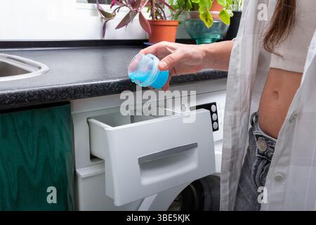 Una giovane donna si trova in una lavatrice a caricamento frontale, tenendo in mano un tappo blu pronto per essere versato, circondata da un pulito spazio di lavanderia con le piante e. Foto Stock