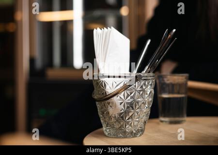 Un dettagliato primo piano di un contenitore di vetro che contiene tovaglioli e cannucce su un tavolo da caffè, abbinato a un bicchiere d'acqua trasparente. Foto Stock