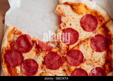 Dall'alto, uno shot ravvicinato di una pizza al peperoni parzialmente mangiata con formaggio fuso e bordi croccanti, appoggiata su carta pergamena. Foto Stock