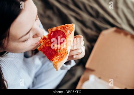 L'immagine dall'alto di una donna assaggia ogni boccone di una fetta di pizza al peperoni mentre si rilassa in un letto a righe. L'istantanea cattura il momento di indulgenza Foto Stock