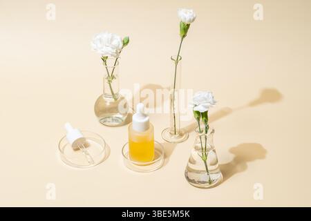 Una bottiglia di contagocce giallo brillante è elegantemente disposta tra vasi in vetro da laboratorio che reggono garofani bianchi, a simboleggiare l'armonia della scienc Foto Stock