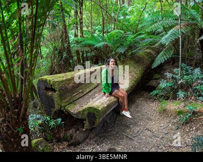 Una donna con una giacca verde siede su un grande tronco di albero caduto circondato da lussureggiante vegetazione della foresta nella Great Ocean Road, Australia. L'impostazione serena Foto Stock