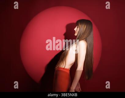 La vista laterale di una giovane donna vestita con un abito rosso si erge su uno sfondo rosso vibrante, che getta un'ombra nitida e distintiva che crea un effetto drammatico Foto Stock