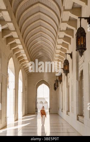 Una donna con un abbigliamento modesto cammina attraverso i corridoi sereni e illuminati dal sole della Moschea del Sultano Qaboos a Mascate, Oman, circondata da archi ornati e hangi Foto Stock