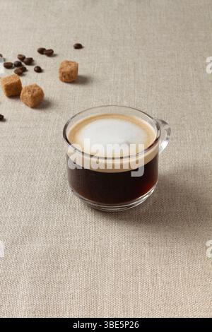 Un espresso macchiato fumante in una tazza di vetro posizionata su una superficie ruvida. Cubetti di zucchero di canna e chicchi di caffè sparsi esaltano l'atmosfera calda e invitante Foto Stock