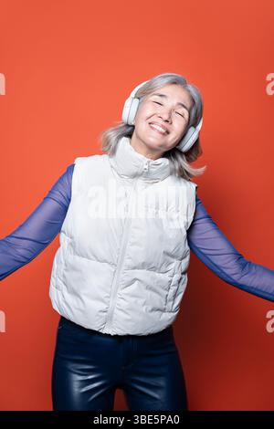 Una donna asiatica anziana ascolta con gioia la musica con le cuffie, indossando un gilet bianco su sfondo arancione. Sorride con gli occhi chiusi, abbracciante Foto Stock