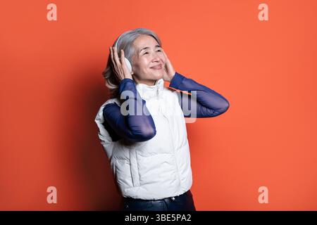 Donna asiatica cinese anziana che indossa le cuffie, sorride e ascolta musica. Vestito con un gilet bianco e maniche blu, appoggiato su un dorso arancione Foto Stock