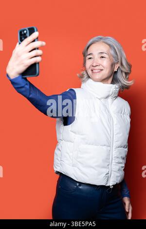 Una donna asiatica cinese senior scatta un allegro selfie con il suo smartphone su un vivace sfondo arancione, indossando un caldo gilet bianco e un contenuto Foto Stock