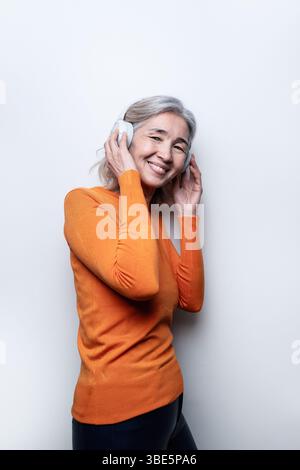Una donna con un maglione arancione ascolta con gioia la musica con le sue cuffie. La sua espressione rilassata e la postura casual creano un'atmosfera allegra e vibrante Foto Stock