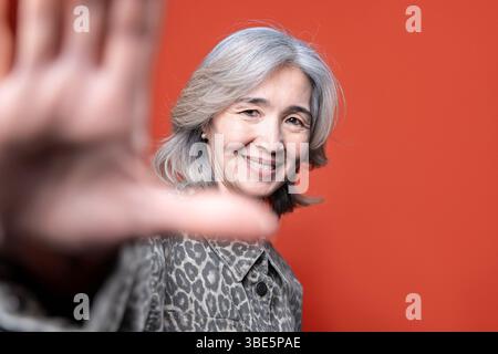 La donna asiatica sorride calorosamente mentre posa di fronte a un vibrante sfondo arancione. I suoi capelli grigi e la sua giacca a motivi aggiungono carattere, trasmettendo la truffa Foto Stock