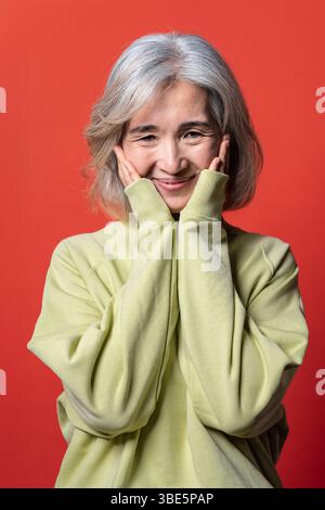 Una gioiosa donna asiatica cinese anziana con i capelli grigi e un maglione giallo posa allegramente su un vibrante sfondo rosso, esprimendo felicità e Foto Stock
