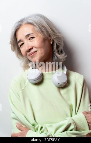 Donna asiatica cinese anziana con capelli grigi, indossa un maglione verde chiaro e cuffie bianche intorno al collo, che guarda con fiducia la fotocamera Foto Stock