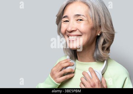 Donna asiatica cinese anziana con i capelli grigi che sorride calorosamente, indossa un maglione verde chiaro, tiene le cuffie bianche e goditi la sua esperienza musicale i Foto Stock