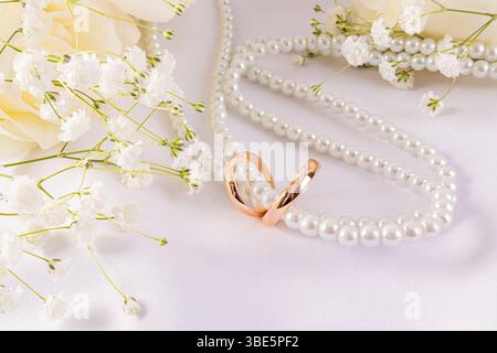 Eleganti fedi da sposa abbinate a delicati fiori e una serie di perle su un morbido sfondo bianco, simboleggiano l'amore e l'unità in un bellissimo seren Foto Stock