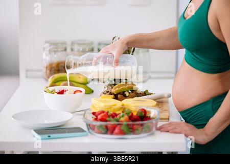 Una donna incinta con un vestito da yoga verde prepara una sana colazione con frutta fresca e latte. L'ambiente enfatizza il benessere e l'alimentazione nutriente Foto Stock