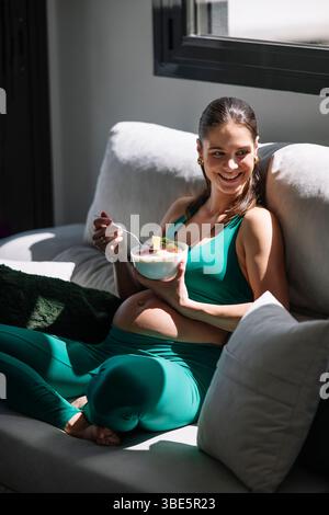 Una donna incinta con un costume da yoga verde si siede su un divano, sorridendo mentre tiene una ciotola di Macedonia di frutta. La luce del sole si diffonde, sottolineando un momento di benessere Foto Stock
