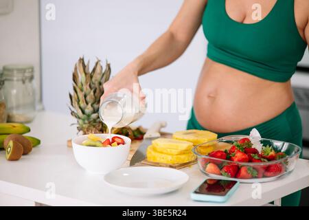Una donna incinta con un vestito da yoga verde prepara una sana colazione con frutta fresca e latte. L'ambiente enfatizza il benessere e l'alimentazione nutriente Foto Stock