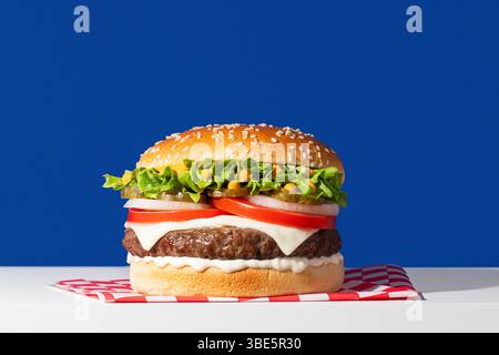 Un delizioso cheeseburger classico presentato su uno sfondo blu. L'hamburger presenta un panino di semi di sesamo, lattuga fresca, fette di pomodoro maturo, cipolla, pic Foto Stock