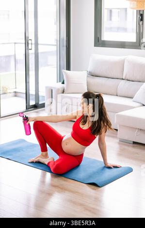 Donna incinta che pratica yoga in un salotto soleggiato mentre indossa un abito rosso, seduta su un tappetino blu da yoga, tenendo in mano una bottiglia d'acqua, promuovendo il benessere Foto Stock