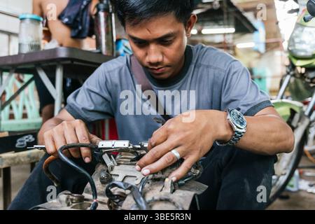Un giovane meccanico si concentra intensamente durante la riparazione del motore di una motocicletta in officina. Le sue mani esperte e la sua espressione mirata mettono in risalto il suo dedic Foto Stock