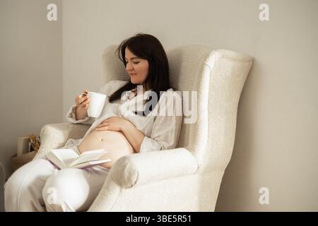Una donna incinta serena si rilassa in una poltrona, sorseggiando un tè e leggendo un libro. Si prepara pacificamente alla maternità, abbracciando il viaggio di Excec Foto Stock