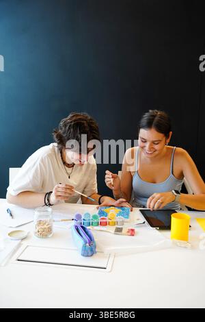 Due adolescenti allegri, un individuo non binario e una donna, si impegnano in un'attività di pittura creativa in una classe. Sono concentrati, condividono una momen Foto Stock