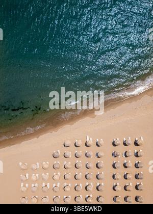 Vista aerea dall'alto verso il basso di una spiaggia sabbiosa con ombrelloni di paglia e lettini allineati in file, onde che lavano delicatamente a riva. Concetto di vacanza estiva Foto Stock