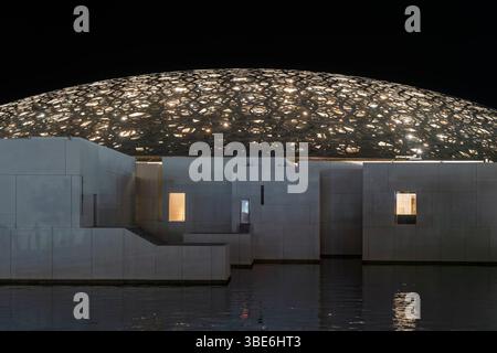 Una vista laterale della cupola del Louvre Abu Dhabi si illumina di luce e si riflette sull'acqua circostante di notte negli Emirati Arabi Uniti. Foto Stock