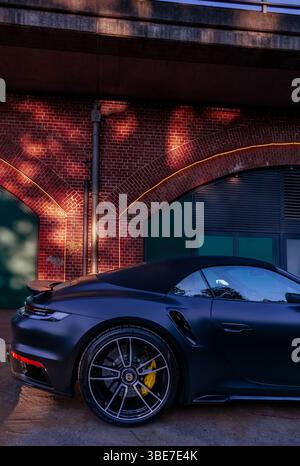 Primo piano di un'auto sportiva Porsche nera parcheggiata di fronte a un edificio ferroviario in mattoni a Berlino, illuminata da una luce soffusa al tramonto e dai riflessi urbani. Foto Stock
