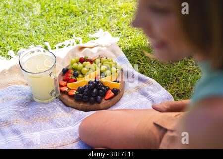 Mostra la donna seduta su una coperta da picnic in erba, con un cartone di legno di frutta e una caraffa di limonata Foto Stock