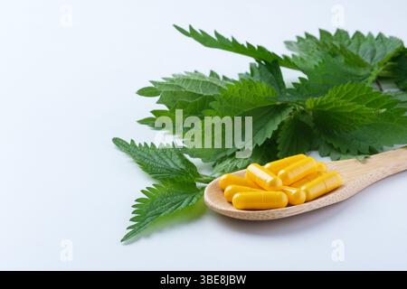 Capsule gialle a base di erbe su un cucchiaio di legno visualizzate accanto a foglie verdi vivaci su una superficie bianca pulita. Ingredienti naturali, medicina alternativa Foto Stock
