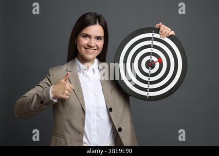 Targeting. Donna d'affari sorridente con freccette e frecce che mostrano i pollici su sfondo grigio Foto Stock