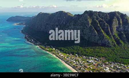 Città costiera sotto le scogliere hawaiane. Vista aerea di una cittadina tropicale sul mare annidata alla base di ripide scogliere verdi lungo il limpido Foto Stock