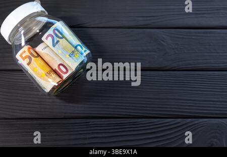Banconote in euro in un vaso di vetro su superficie di legno scuro. Vaso di vetro contenente varie banconote in euro, compresi i tagli da cinquanta, venti e dieci Foto Stock