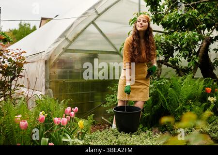Una giovane donna con un velo tende gioiosamente a piante in una vibrante serra mentre indossa i guanti. Foto Stock