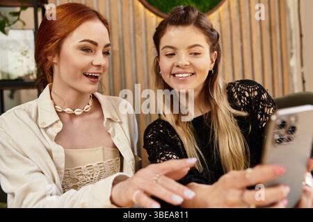 Due amici sorridono e condividono un momento insieme utilizzando uno smartphone in un bar alla moda. Foto Stock