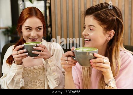 Due giovani donne condividono risate e bevande verdi in un moderno ambiente da caffè, abbracciando l'amicizia. Foto Stock