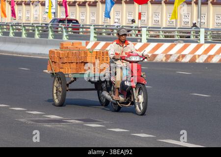 DANANG, VIETNAM, APR 27 2025, Un uomo su una motocicletta che trasporta mattoni con un rimorchio su una strada urbana soleggiata Foto Stock