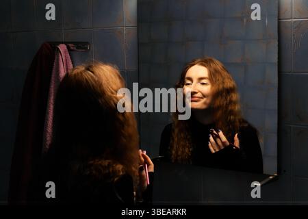 Una giovane donna con lunghi capelli rossi ondulati applica il rossetto in un bagno poco illuminato, il suo riflesso visibile nello specchio. Foto Stock