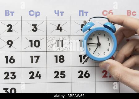 Una mano trattiene una piccola sveglia, in background è un calendario con il giorno segnato e la parola vacanza Foto Stock