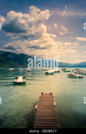 Un pontile in legno si estende sulle tranquille acque del lago Annecy, circondato da montagne e punteggiato di barche colorate. Alta Savoia . Francia Foto Stock