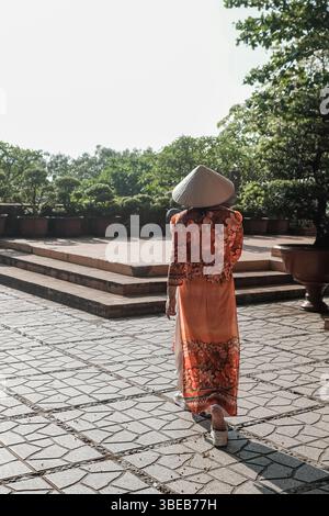 Signora vietnamita con abito tradizionale Ao dai Vietnam. Vista posteriore di una bella donna asiatica che viaggia in abiti tradizionali e cappello conico in un giardino Foto Stock